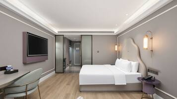 A hotel room with a bed, bedside table, TV, desk, chair, and a bathroom visible through a door.