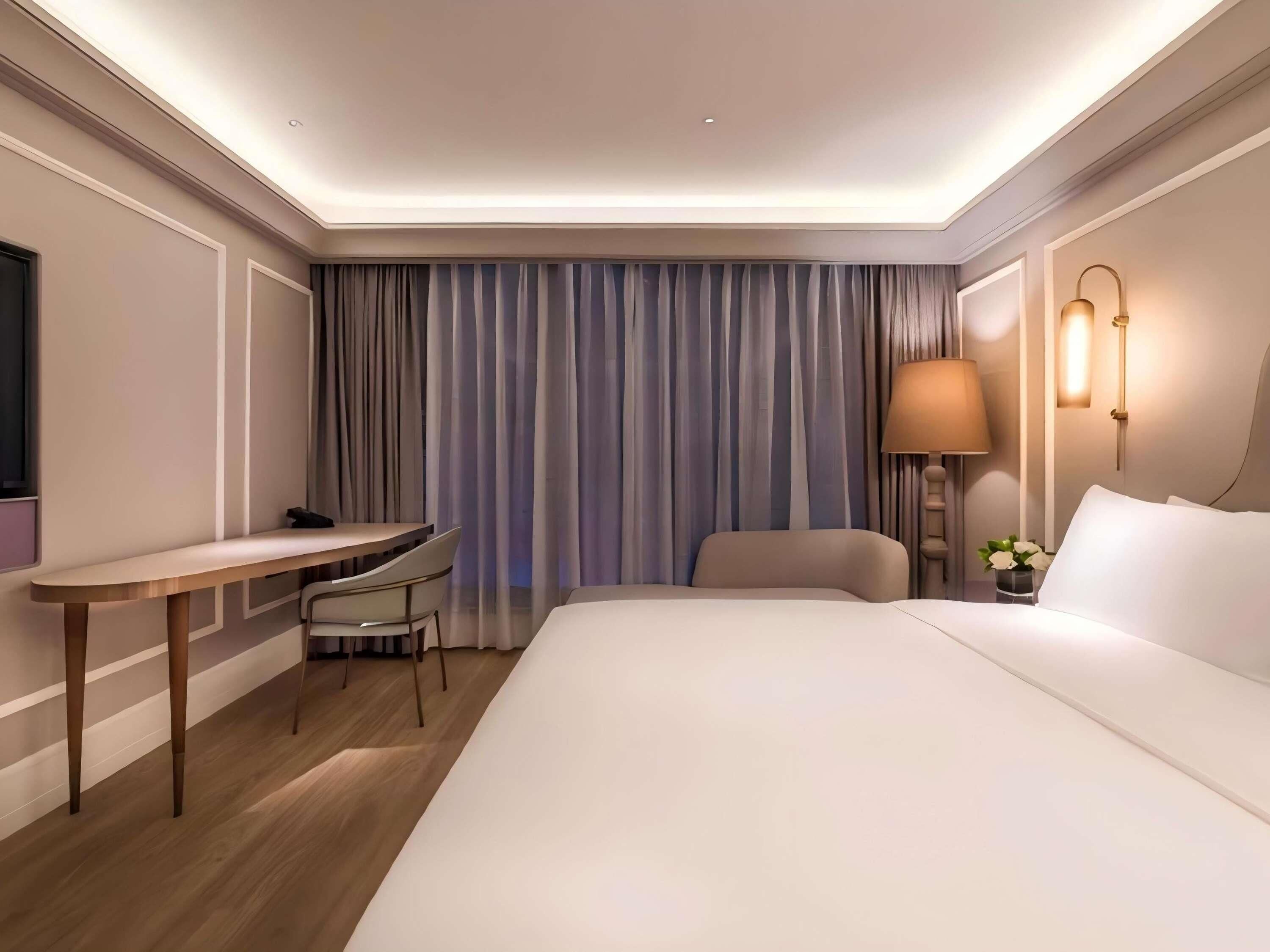 Una habitación de hotel con cama, escritorio, silla, lámpara y cortinas.