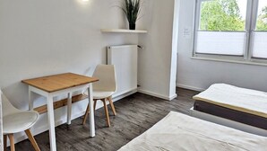 A small, clean room with a wooden table, two white chairs, a bed, a radiator, and a window with a view of greenery. - Gästehaus Koblenz (Koblenz)