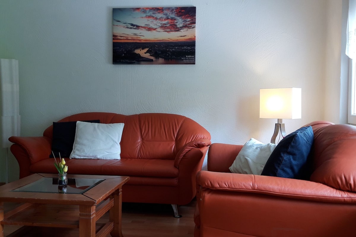A living room with a red leather sofa, a glass coffee table, and a painting of a sunset on the wall.