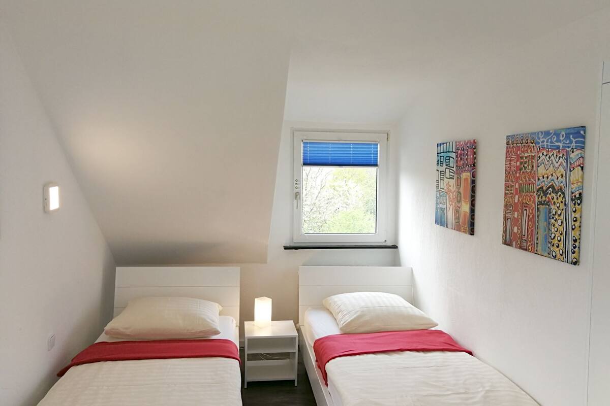 A small, bright room with two beds, a window with blinds, and colorful artwork on the walls.