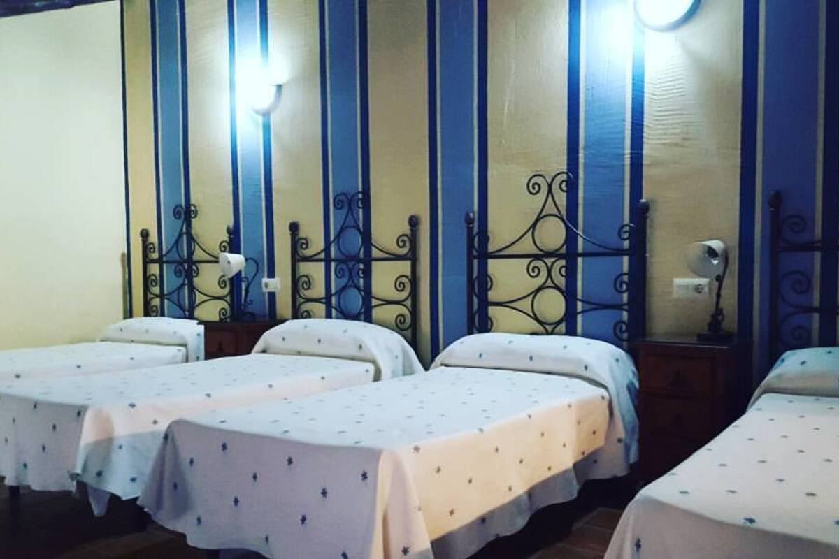 A room with four single beds, each with a white bedspread and a small decorative pillow. The room has a simple wooden floor and walls painted in a two-tone color scheme.