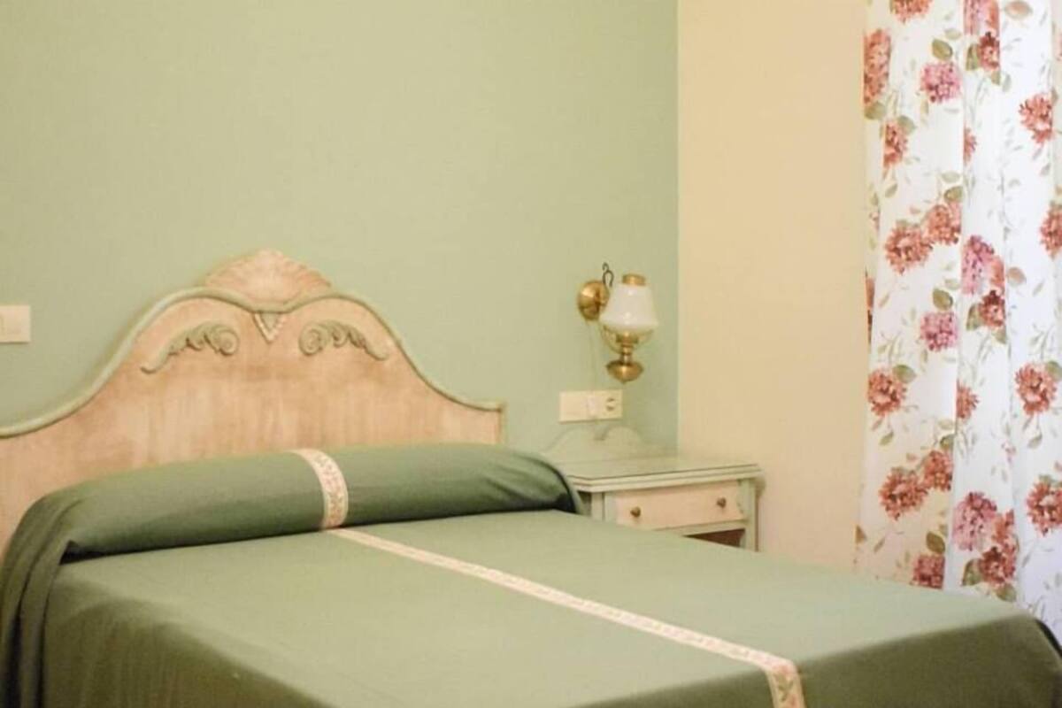 A bed with a wooden frame and green bedding, a nightstand with a lamp, and a floral curtain.