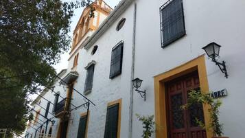 A white building with a yellow door and windows with black shutters. There are potted plants and a metal gate in front.