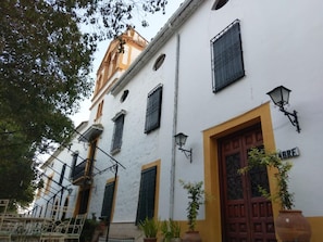 A white building with a yellow door and windows with black shutters. There are potted plants and a metal gate in front. - Casa Rural Herrera (Villacarrillo)