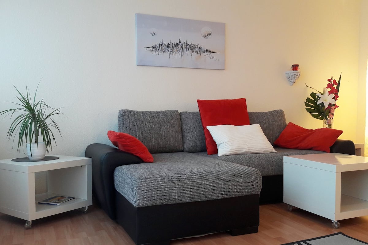 A modern living room with a grey sofa, red and white cushions, a small side table with a plant, and a white side table with a lamp.
