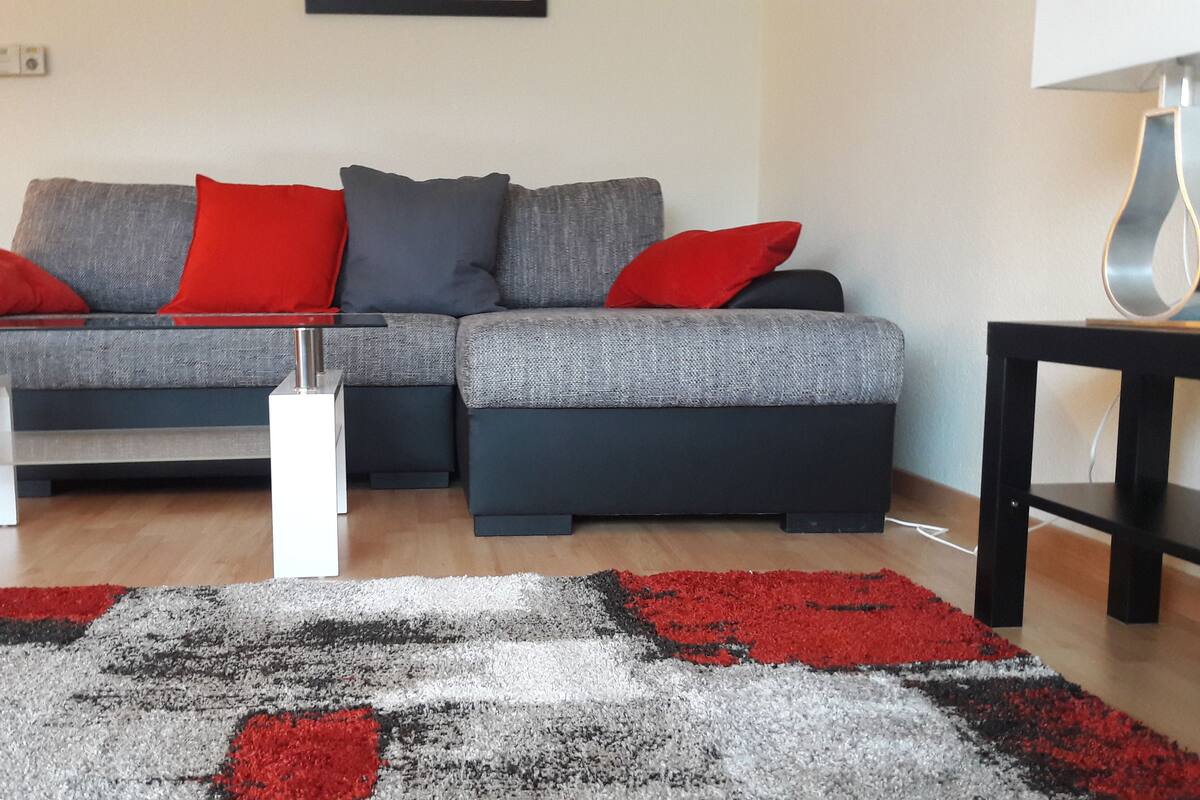 A modern living room with a grey sofa, red and grey cushions, a glass coffee table, and a black side table.