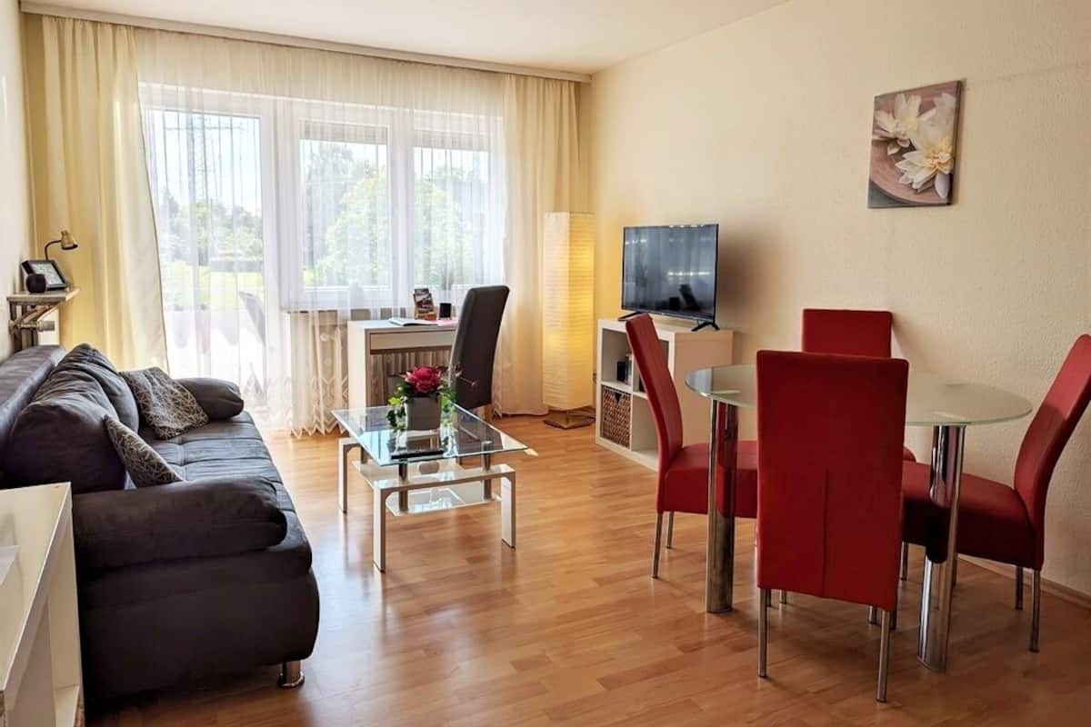 A modern living room with a sofa, a glass coffee table, and a dining area with red chairs and a glass table.