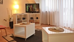 A modern living room with a television, white furniture, and a large window with sheer curtains. - Apartmenthaus Abendsonne (Koblenz)