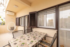 Ein Balkon mit Tisch, der mit einem Tischset mit Pflanzenmuster bedeckt ist, einem Hängesessel und einem Fenster mit Fensterläden.