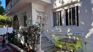 A white building with a balcony, a small garden with a tree and flowers, and a table with two chairs outside.