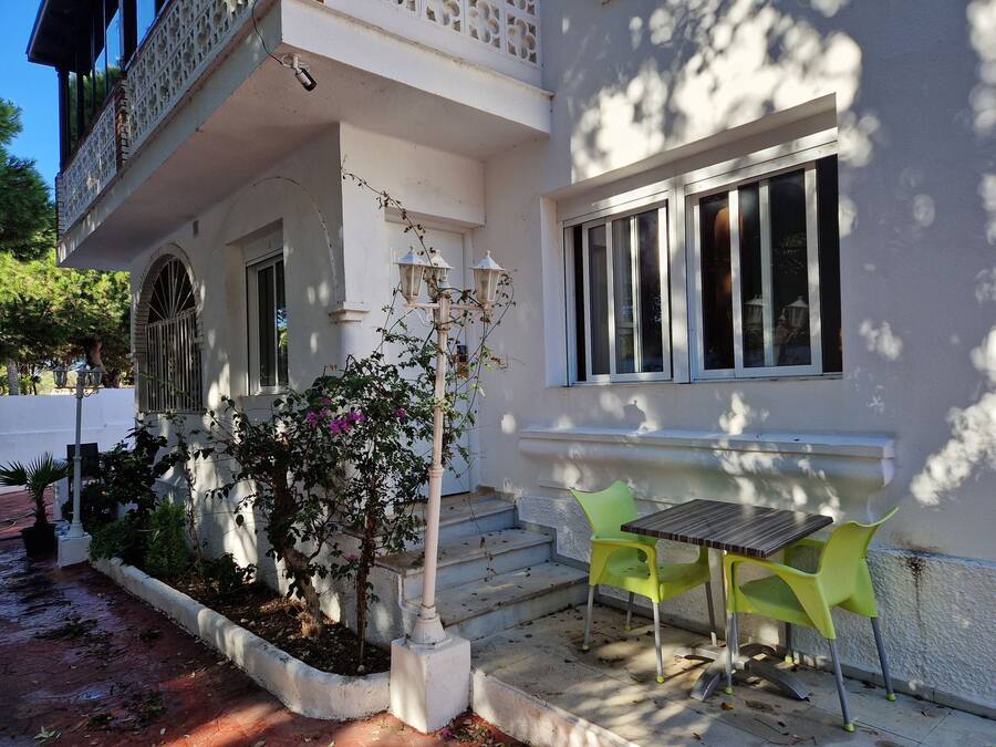 A white building with a balcony, a small garden with a tree and flowers, and a table with two chairs outside.