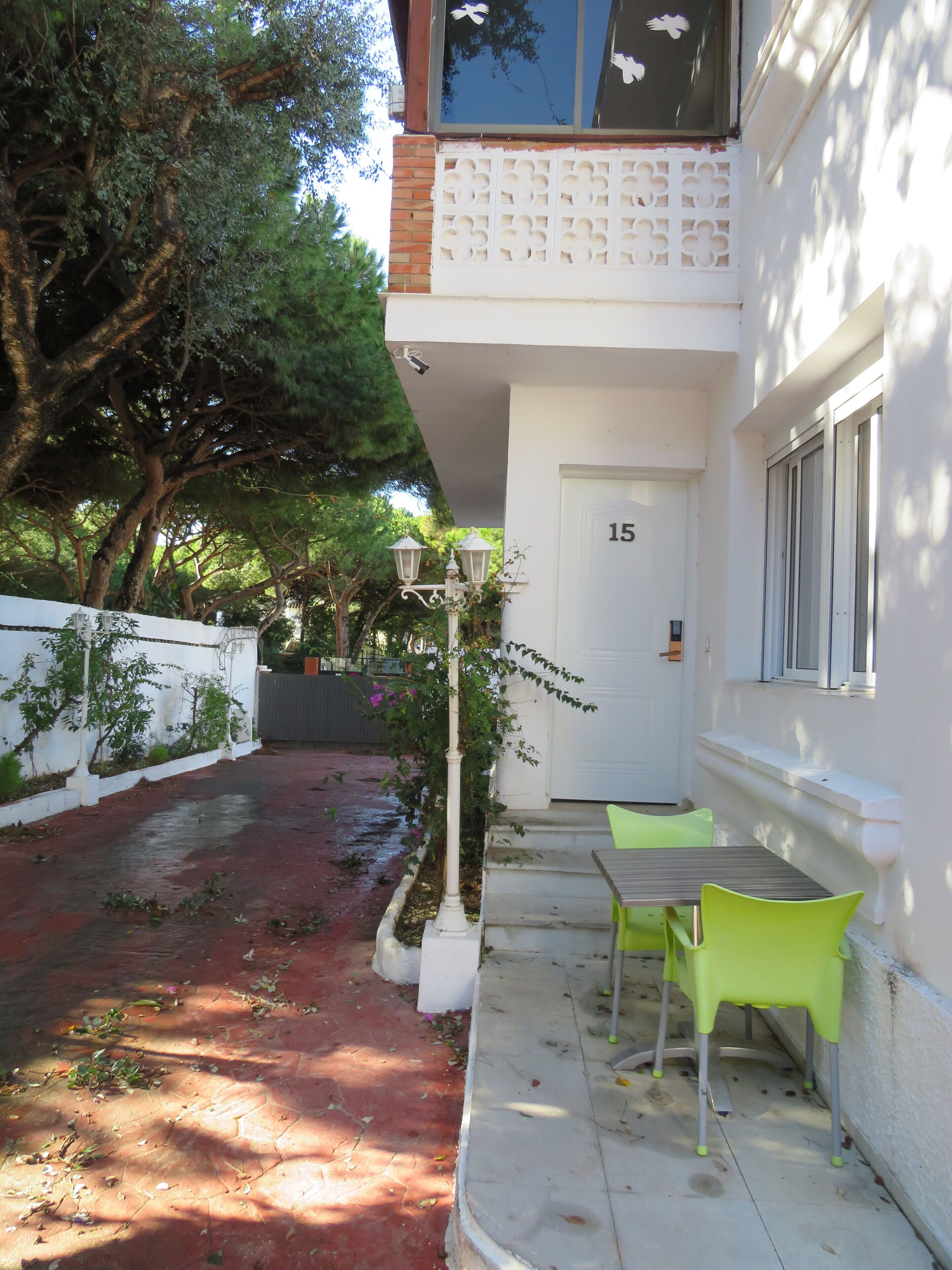 Un edificio con fachada blanca, una puerta con el número 15 y un pequeño espacio exterior con sillas verdes.