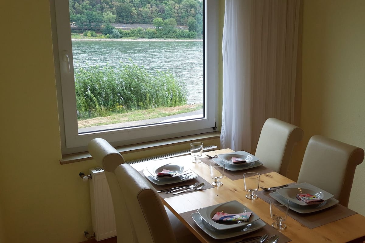 Ein Esstisch mit Stühlen, Tellern, Besteck und Gläsern, gedeckt für eine Mahlzeit. Ein Fenster bietet Blick auf einen Fluss und viel Grün.