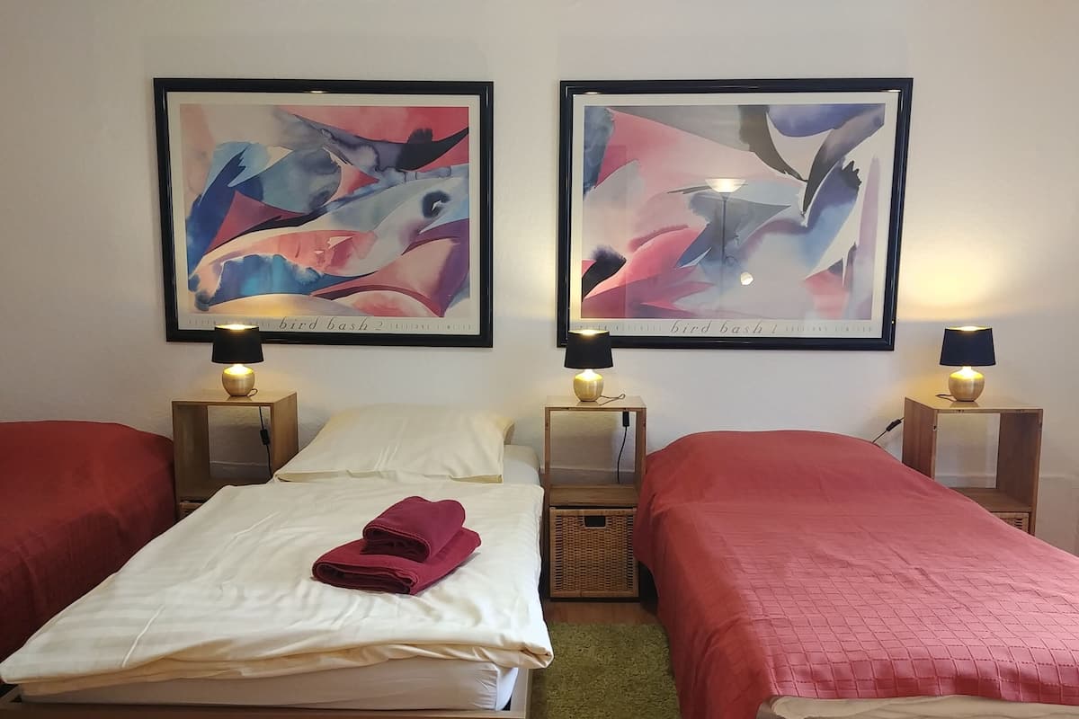 Ein Hotelzimmer mit zwei Betten, jeweils mit roter Bettdecke und weißen Laken. Darüber hängen zwei gerahmte abstrakte Gemälde, und jedes Bett hat einen kleinen Nachttisch mit einer Lampe.