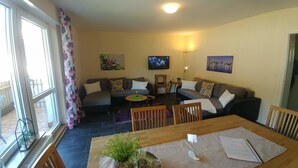 A living room with a sofa set, a dining table with chairs, a TV mounted on the wall, and a balcony with a view. - Ferienwohnung Koblenz City (Koblenz)