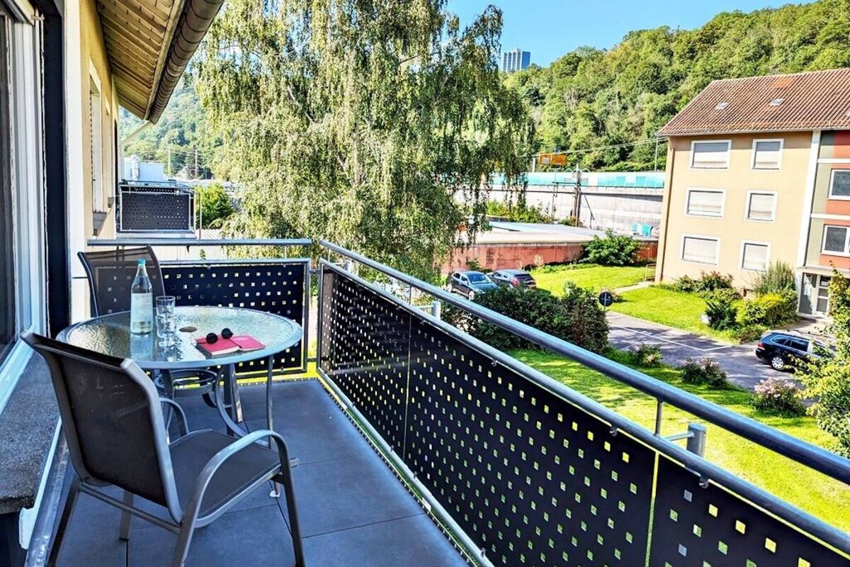 Un balcon avec une table et des chaises en verre, donnant sur un paysage verdoyant et des bâtiments.