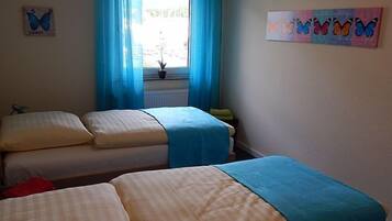 A room with two beds, a window with blue curtains, a wall with a butterfly painting, and a small plant on the windowsill.