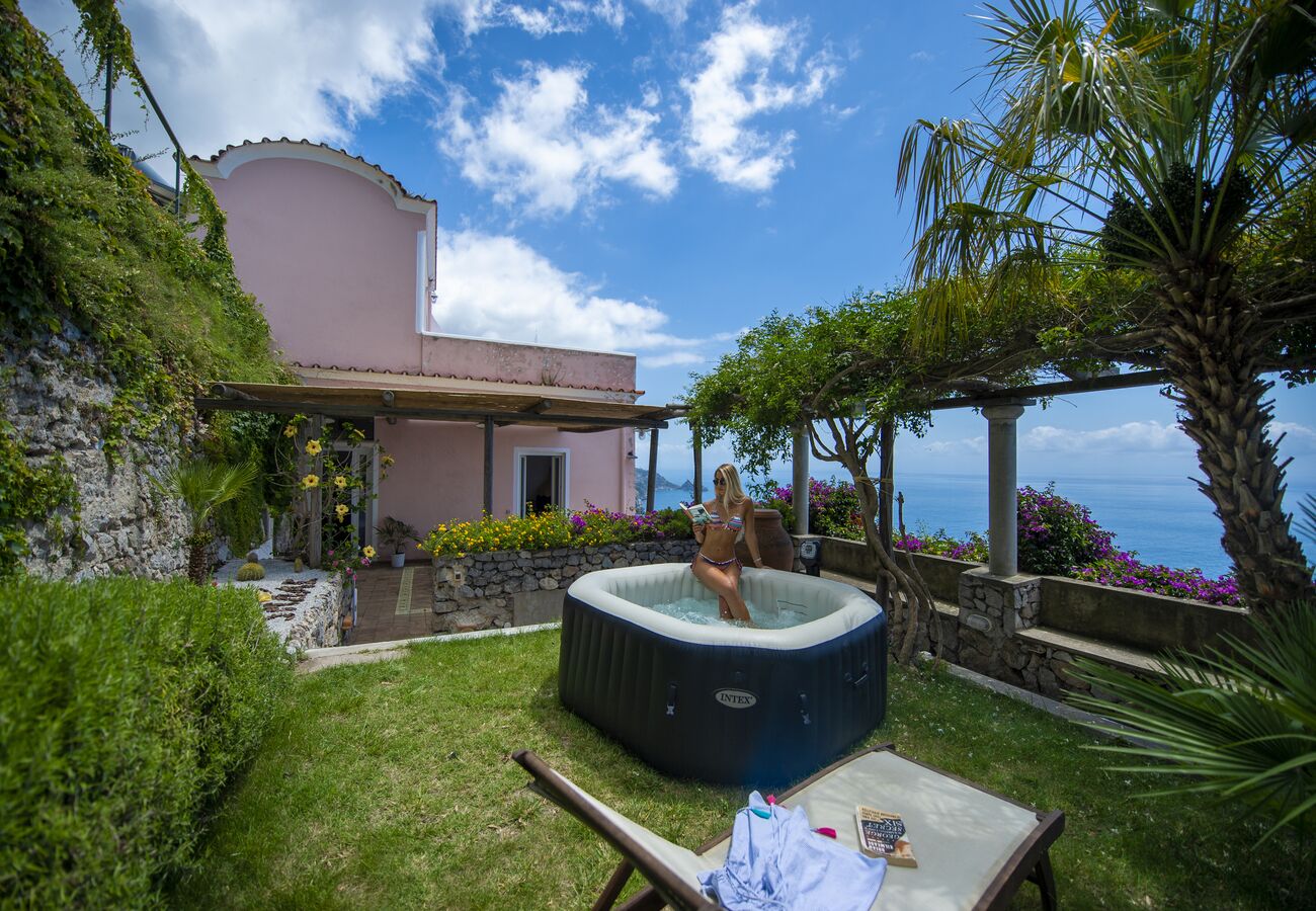 A hot tub in a garden with a view of the sea, surrounded by greenery and a pink building.
