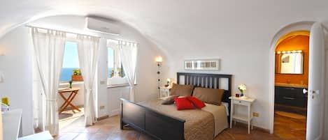 A bedroom with a bed, bedside tables, a small table with a chair, a window with curtains, and a view of the sea.