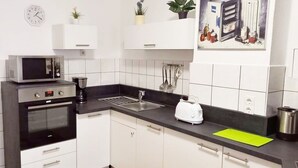 Een moderne keuken met witte kastjes, een zwart aanrecht, een magnetron, een oven, een broodrooster en een spoelbak. Er staan planten op de kastjes en een klok hangt aan de muur.
