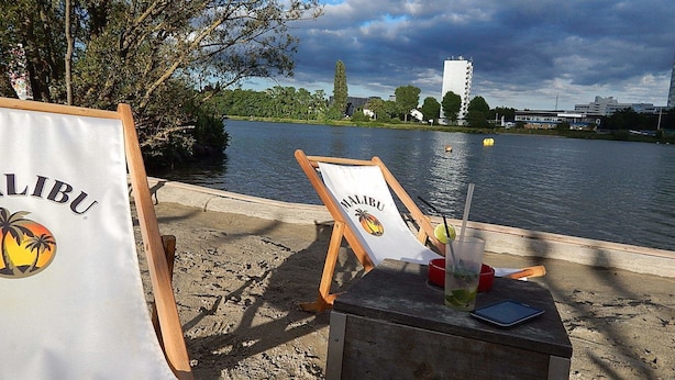 Een strandsetting met twee loungestoelen, voorzien van de naam 'Malibu', en een houten tafel met drankjes en een tablet.
