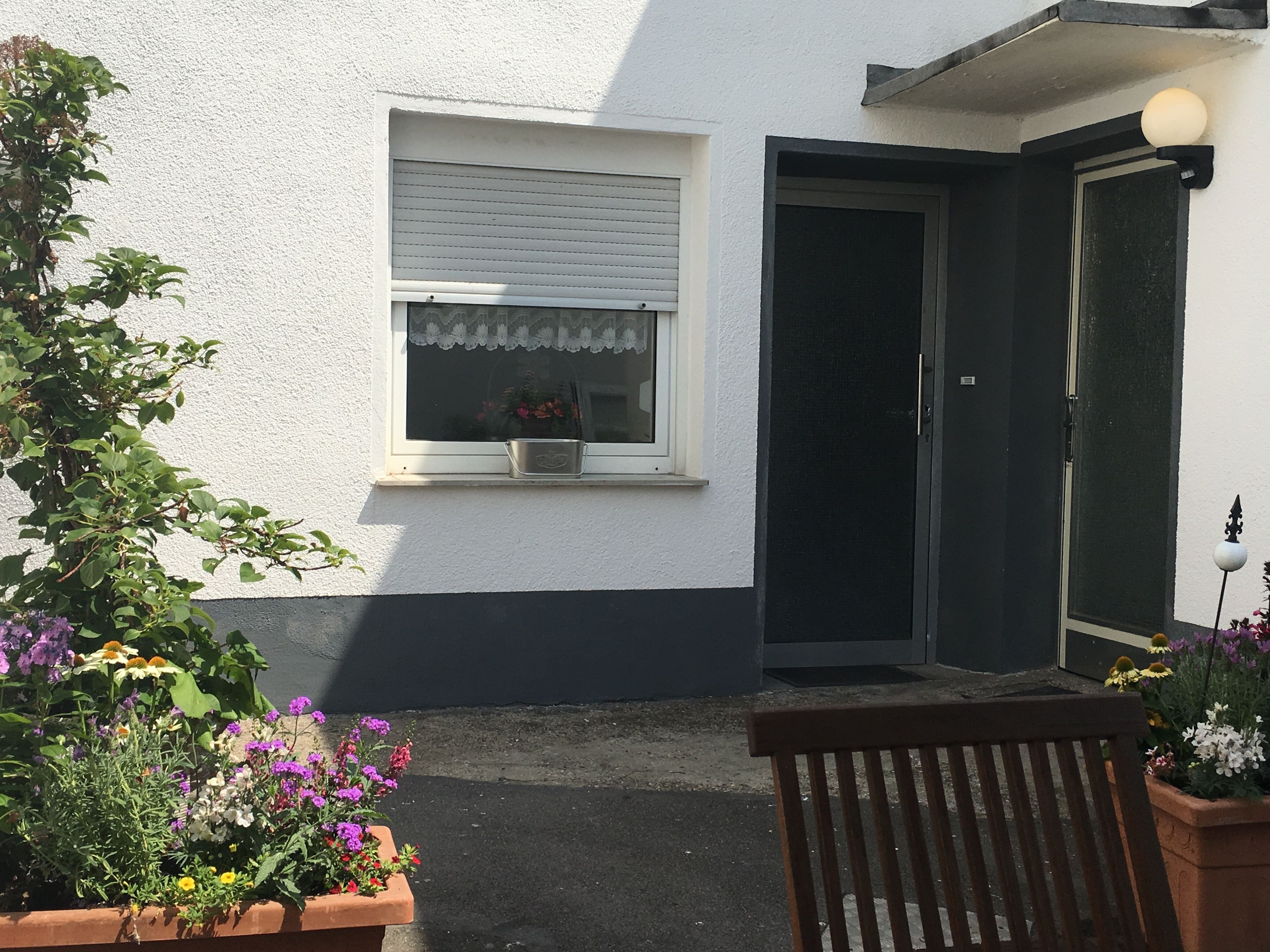 A small house with a white exterior, a window with a white frame, and a door with a black frame. There is a bench in front of the house and a potted plant with flowers to the left.