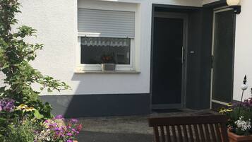 A small house with a white exterior, a window with a white frame, and a door with a black frame. There is a bench in front of the house and a potted plant with flowers to the left.