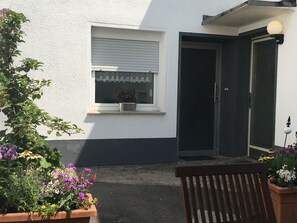 A small house with a white exterior, a window with a white frame, and a door with a black frame. There is a bench in front of the house and a potted plant with flowers to the left.