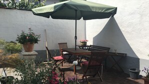 Een terras buiten met een tafel en stoelen, omringd door potplanten en een groene parasol.