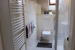 A small bathroom with a toilet, a towel rack with towels, a shower, and a toilet paper holder.