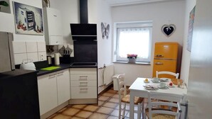 Een compacte keuken met witte kastjes, een zwarte afzuigkap, een kleine eettafel met stoelen, en een vintage oranje koelkast.
