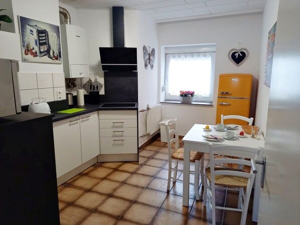 A compact kitchen with white cabinets, a black range hood, a small dining table with chairs, and a vintage orange refrigerator.