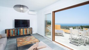 A modern living room with a flat-screen TV, a wooden cabinet, and a coffee table. It features a large sliding glass door leading to an outdoor seating area with a view of the sea. - Miramar by Interhome (Moraira)