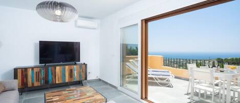 A modern living room with a flat-screen TV, a wooden cabinet, and a coffee table. It features a large sliding glass door leading to an outdoor seating area with a view of the sea.