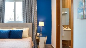 Quarto de hotel com cama, mesa de cabeceira com abajur, banheiro com espelho e vista para a cidade pela janela.