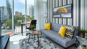 Uma sala de estar moderna com um sofá cinzento, almofadas amarelas e pretas, um tapete preto e branco, uma mesa de centro e um quadro com uma imagem de uma árvore.