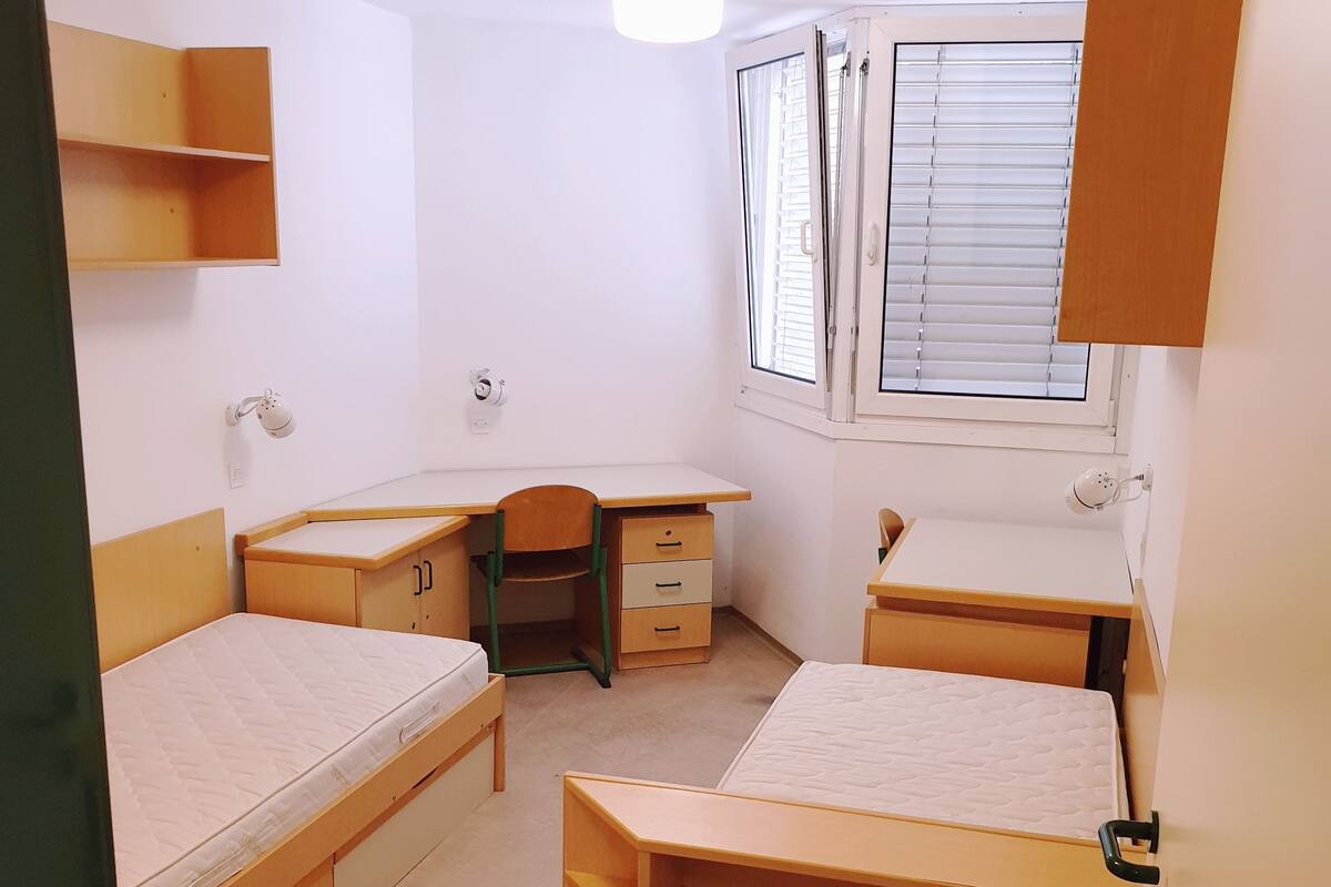 A compact room with two beds, a desk, and a window with blinds.