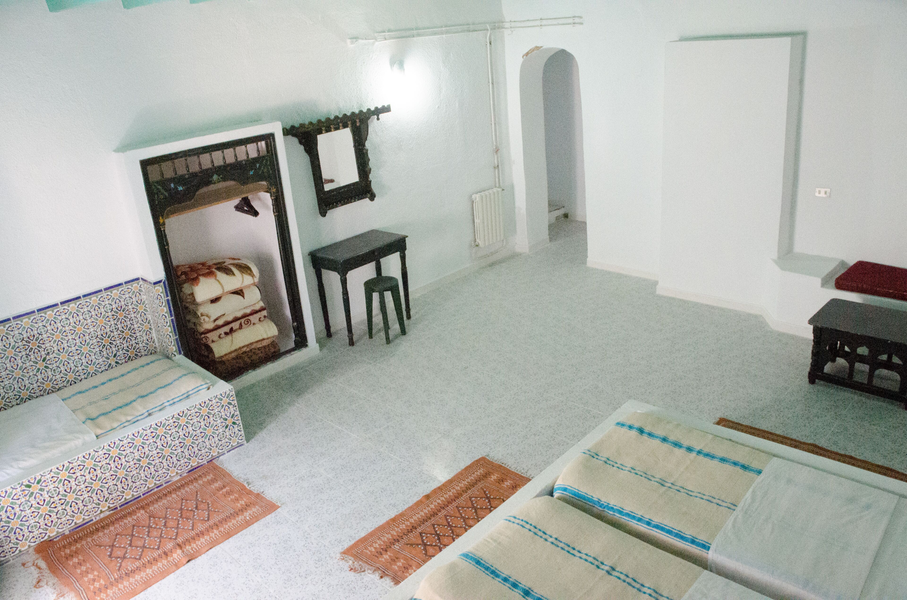 A room with a bed, a small table, a chair, a mirror, and a radiator.