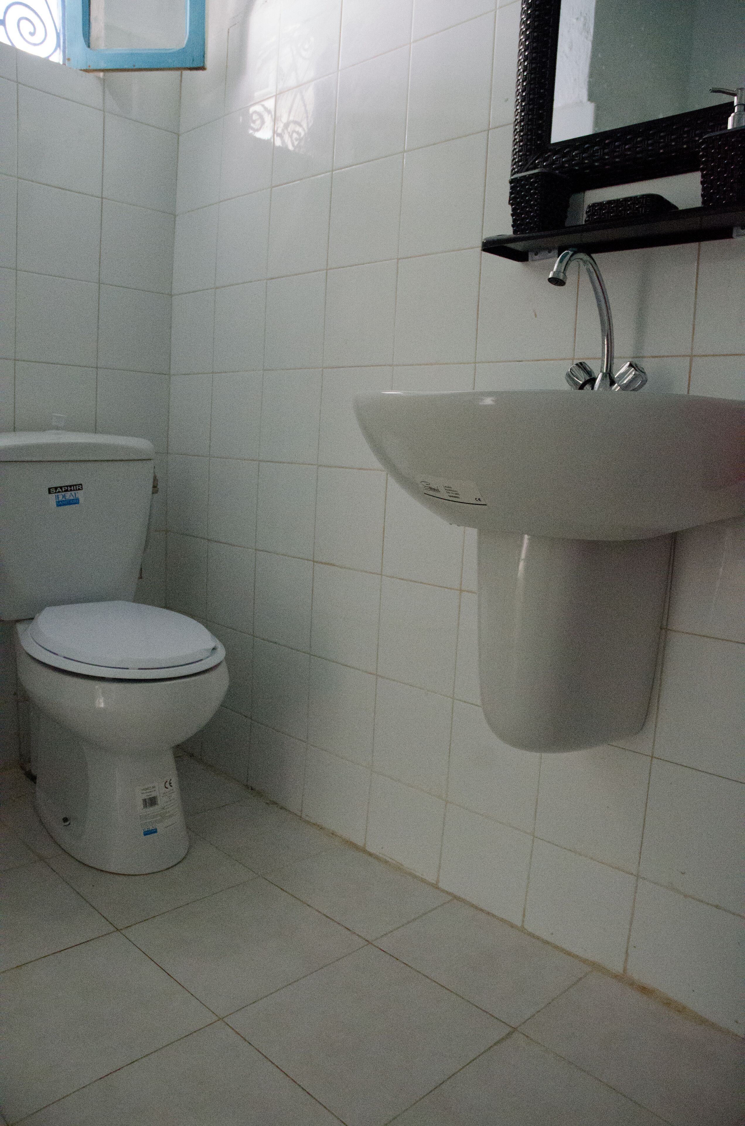 Une salle de bain avec des toilettes et un lavabo blancs, des murs recouverts de carreaux blancs et un miroir à cadre noir au-dessus du lavabo.