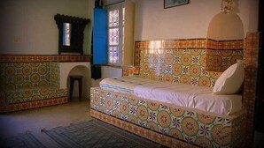 Une pièce avec un lit à motifs, un petit tabouret, une fenêtre avec un rideau et une lampe fixée au mur.