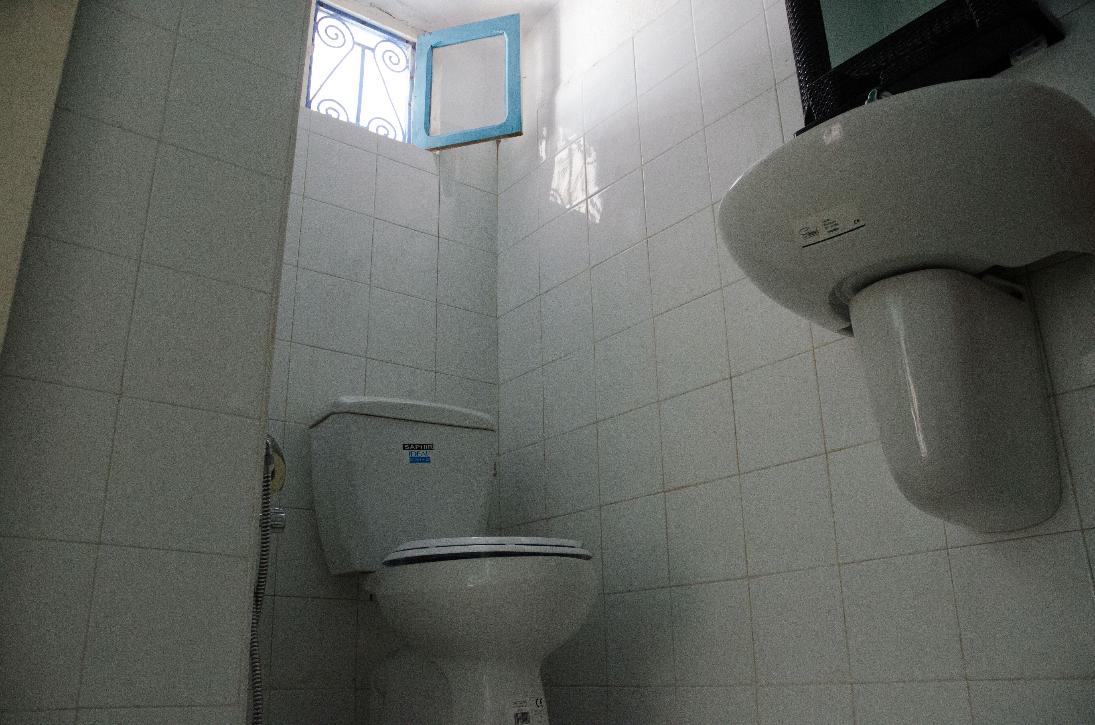 Une salle de bain avec des murs recouverts de carreaux blancs, des toilettes, une douche et une fenêtre à cadre bleu.