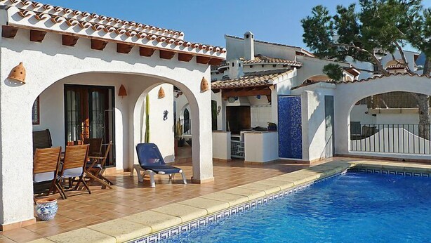 A poolside area with a tiled deck, a covered seating area with chairs, and a pool with a blue border. - Esperanza by Interhome (Pego)