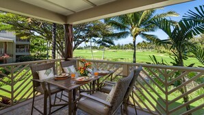 A covered outdoor dining area with wicker furniture and a table set for two, overlooking a well-manicured lawn and palm trees. - 🏖️Family-Friendly | Pools, Beach Gear, Golf & Walk to A-Bay Adventures (Waikoloa)