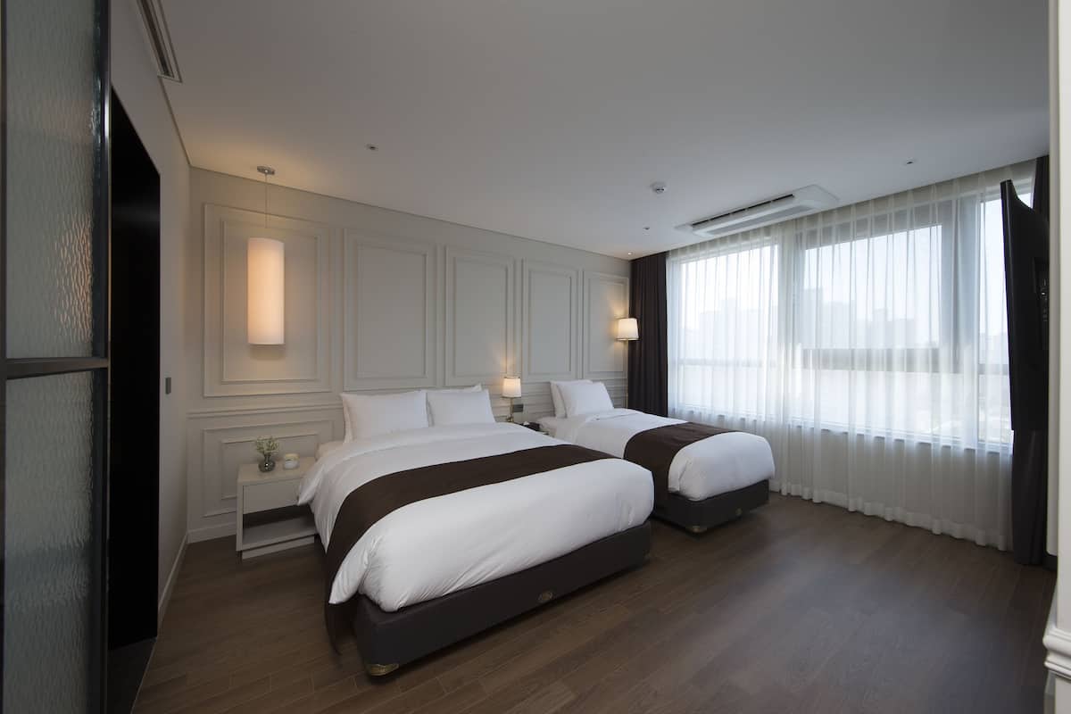 A hotel room with two beds, a nightstand, a lamp, and a large window with sheer curtains.