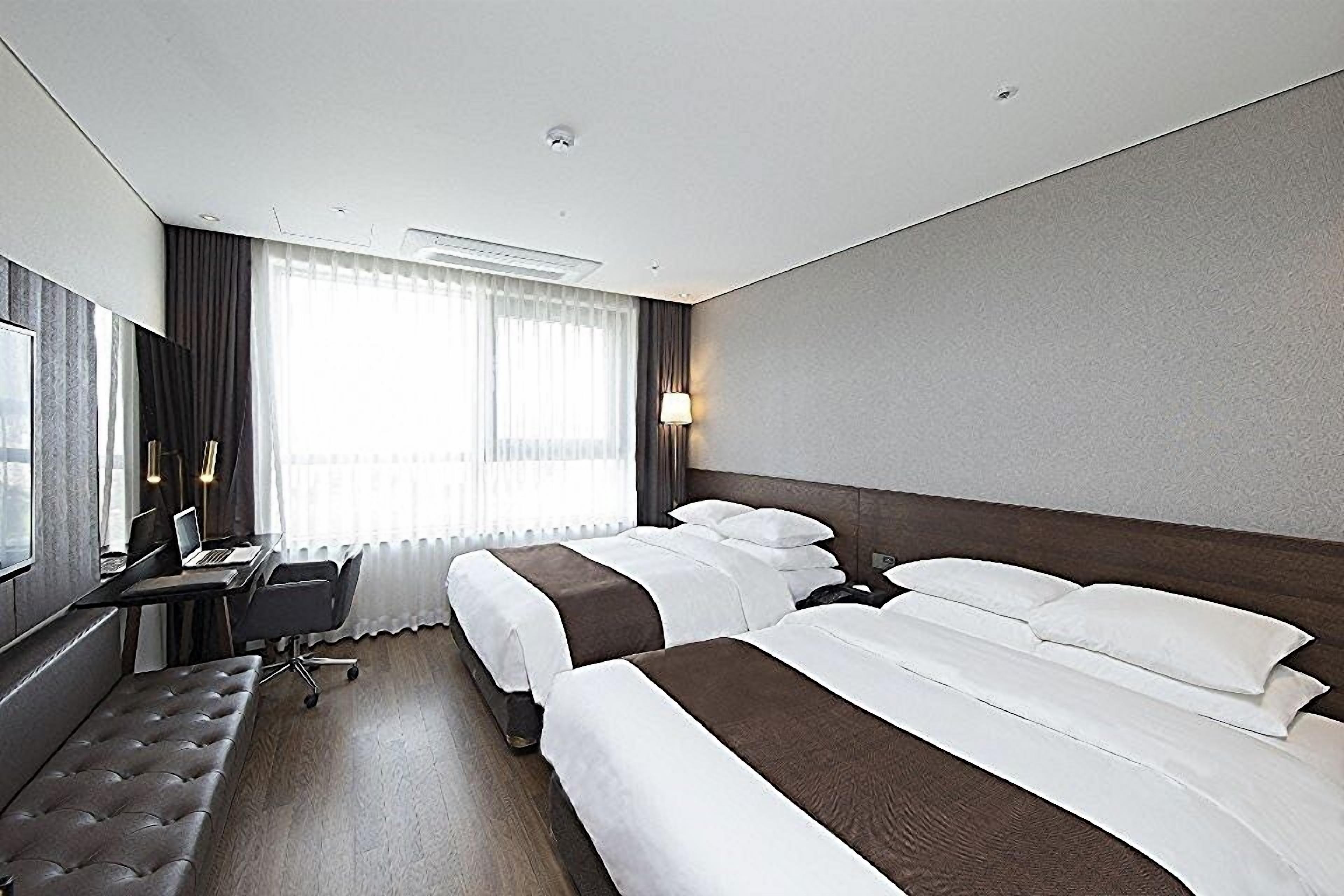 A hotel room with two beds, a desk, a chair, and a large window with curtains.