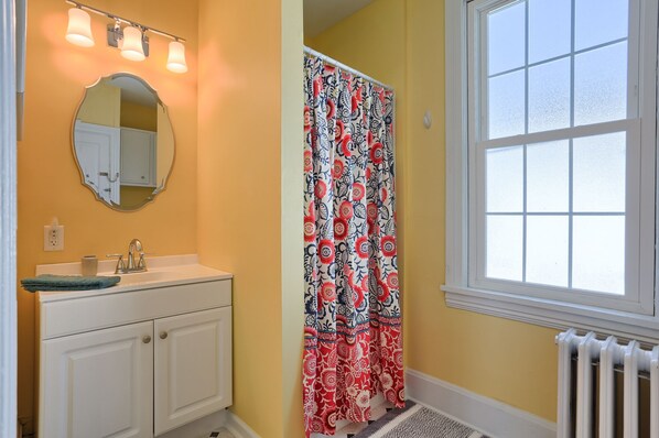 A bathroom with a yellow wall, a white sink with a mirror above it, a patterned shower curtain, and a window with a view of the sky.