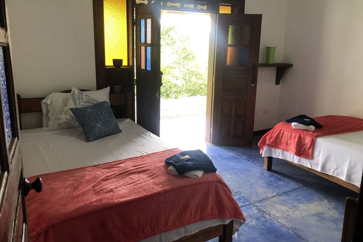 Een kamer met twee bedden, elk met een rode sprei, een houten hoofdbord en een witte matras. Er is een houten deur met ruitpanelen en een klein plankje erboven.