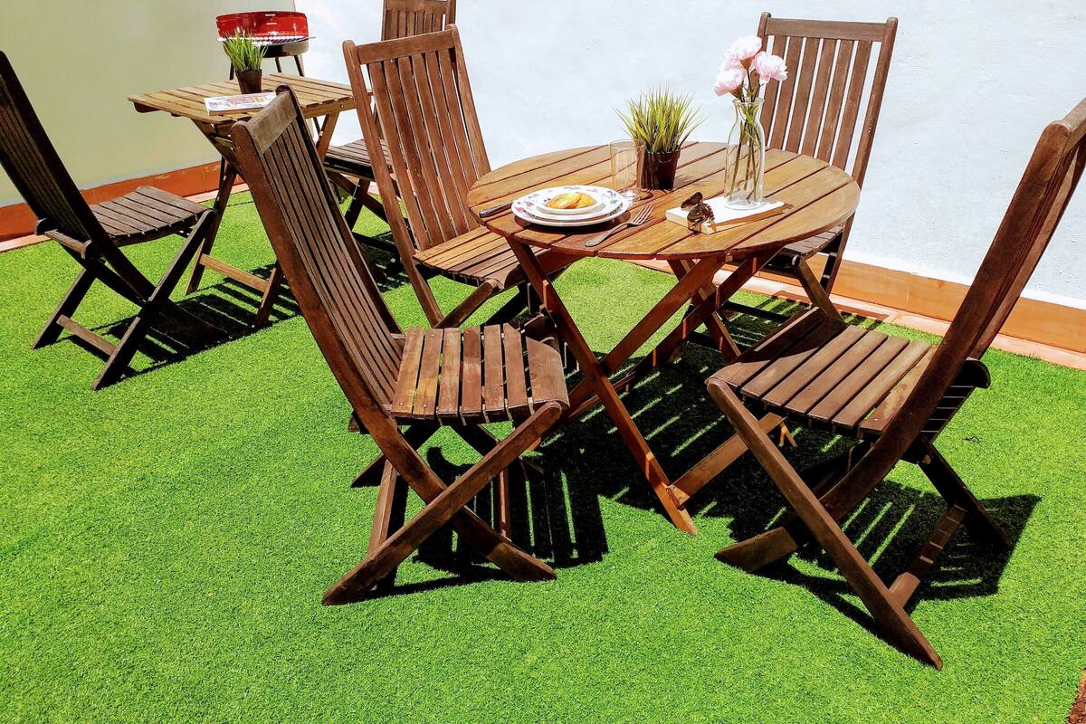 A wooden table and chairs set on artificial grass with a white wall in the background.