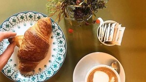 A croissant on a patterned plate, a cup of coffee with latte art, and a small plant with decorative elements. - Bayard101 (Naples)